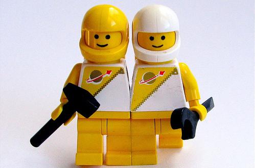 Two LEGO minifigures dressed in white and yellow spacesuits with the classic space logo, one holding a wrench and the other a walkie-talkie.