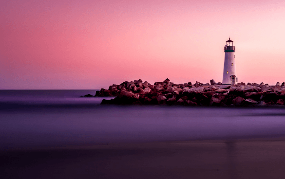 A lighthouse on a rocky breakwater with a gradient pink and purple sky, likely at sunrise or sunset, with smooth water in the foreground due to a long exposure effect.