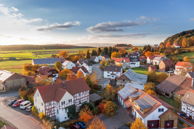 Aerial view of a small village with traditional timber-framed houses surrounded by autumn-colored trees, fields, and a distant forest under a partly cloudy sky with a warm glow from the sun.