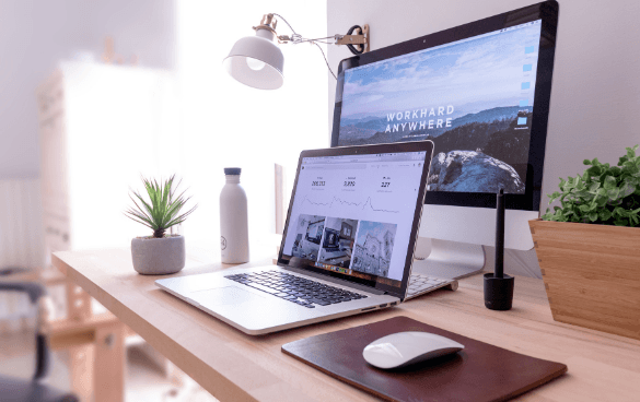 A tidy workspace with a laptop displaying a website design, a desktop monitor with inspirational text, a desk lamp, two potted plants, and a water bottle on a wooden desk.