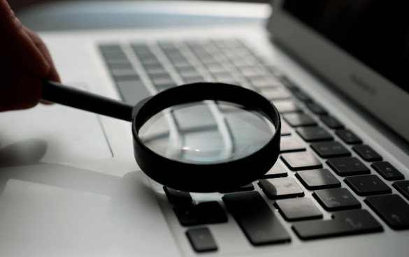 A person's hand holding a magnifying glass over a laptop keyboard, magnifying the keys.