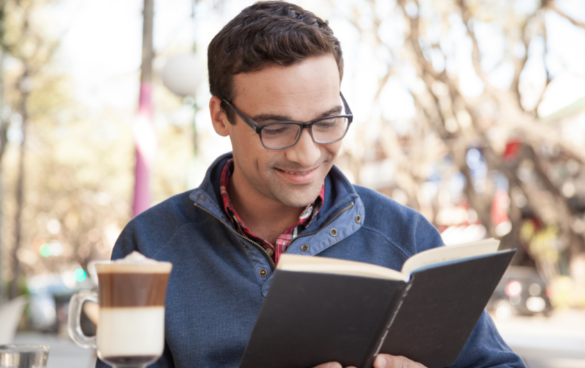 A man smiles as he reads a book next to a glass of hot chocolate