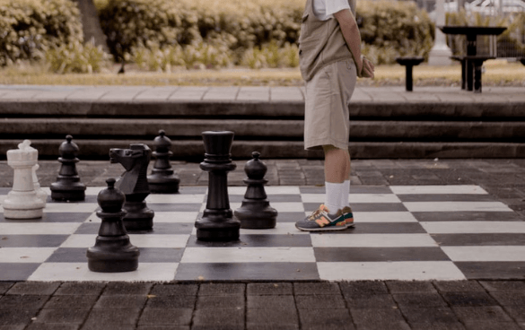 A person stands on an oversized outdoor chessboard with large chess pieces, dressed in shorts and multicolored sneakers, in a park setting.