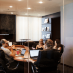 Four people in a business meeting inside a modern office with glass walls, laptops on the conference table, and a flat-screen monitor in the background.