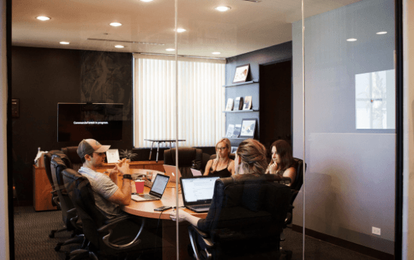 Four people in a business meeting inside a modern office with glass walls, laptops on the conference table, and a flat-screen monitor in the background.