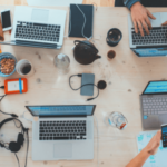 Overhead view of a busy workspace with multiple people using laptops, smartphones, and taking notes, surrounded by personal items like headphones and coffee cups on a wooden table.