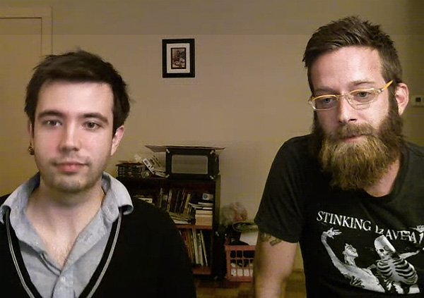 Two men in front of a bookshelf, one with short dark hair and a black shirt, the other with a beard, glasses, and a graphic t-shirt, looking at the camera.