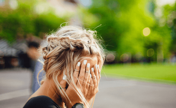 Person with braided blonde hair covering their face with hands, set against a blurred green outdoor background.