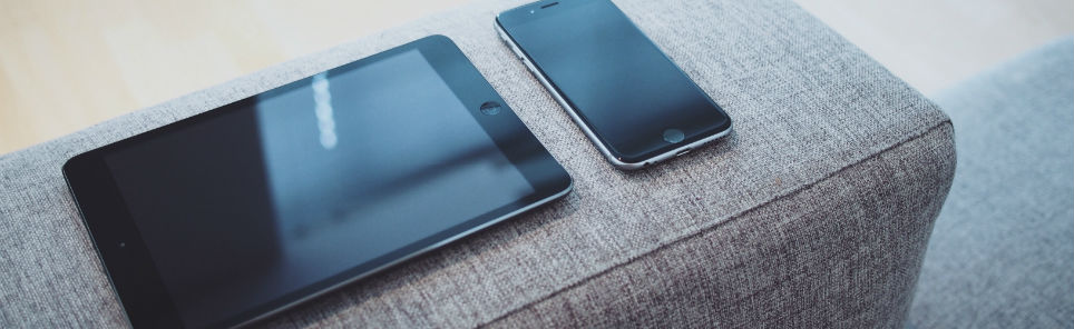 A tablet and a smartphone with black screens lying on a gray fabric ottoman.