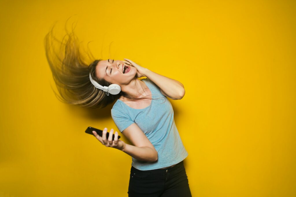 Joyful woman with headphones holding a smartphone, hair tossed to the side, against a yellow background, expressing happiness as if singing or dancing.