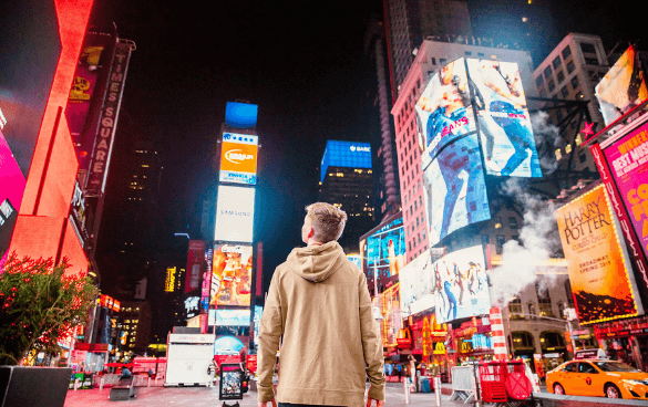 A person from behind observing the vibrant and illuminated billboards of Times Square at night, with a yellow taxi and other vehicles on the street.