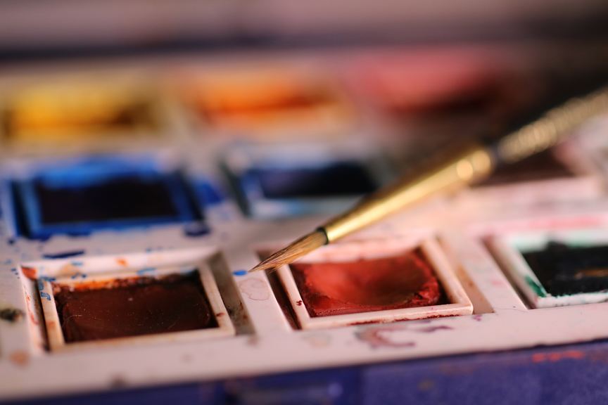 Close-up of a used watercolor paint set with a paintbrush resting on top.