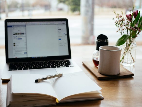 A cozy workspace with an open laptop, notebook with a pen, a white mug on a tray, a glass bottle with dark liquid, and a vase with fresh flowers on a wooden table.