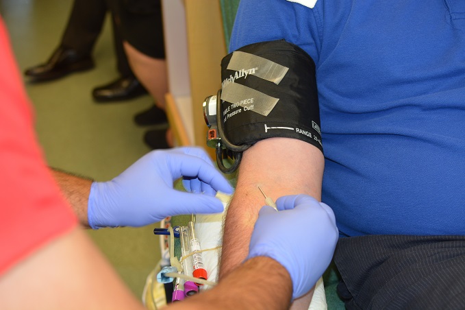 A healthcare professional wearing blue gloves is performing a blood draw on a patient's arm, which has a blood pressure cuff, with multiple vacutainer tubes ready for sample collection.