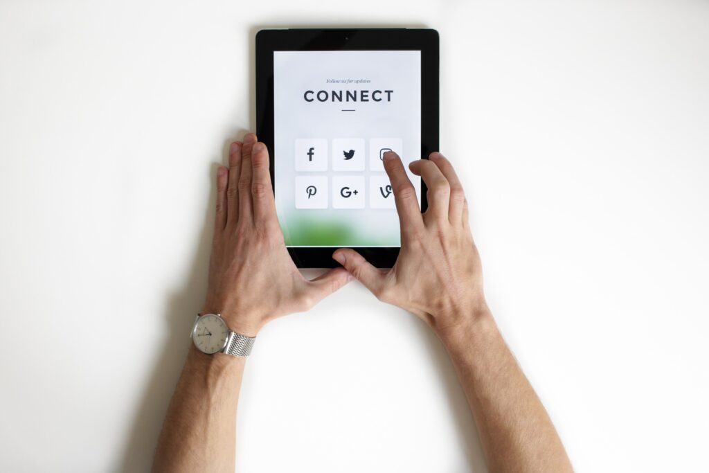 Person holding a tablet displaying social media connect screen with icons for Facebook, Twitter, Pinterest, Google Plus, and a link symbol, against a white background, wearing a silver mesh band wristwatch.