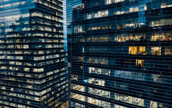 Close-up of modern office buildings with reflective glass facades, showing a pattern of illuminated and non-illuminated windows during twilight.