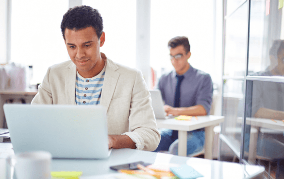 Man typing on a computer with another man on a computer behind him