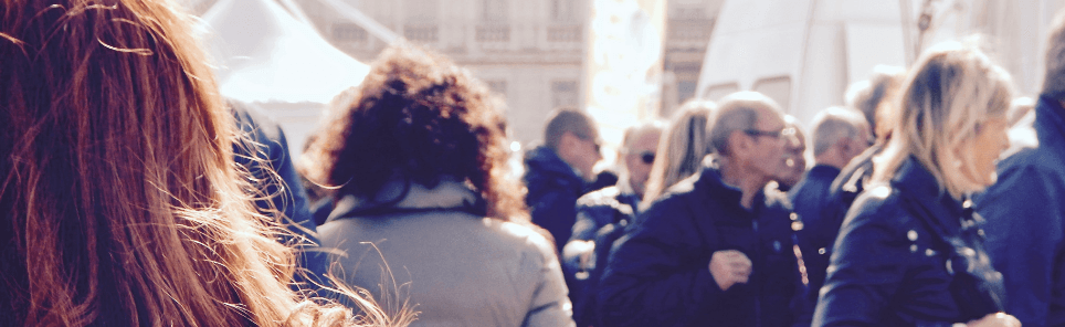 A crowd of people gathered outdoors, with the focus on the back of two individuals' heads, one with reddish hair and the other with curly hair, on a sunny day.