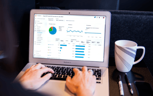 Person using a MacBook Air displaying analytics data, with a coffee cup, pens, and a notebook on the table.