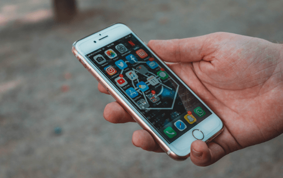 A person's hand holding a rose gold iPhone with the screen displaying colorful app icons, with a blurred outdoor background.