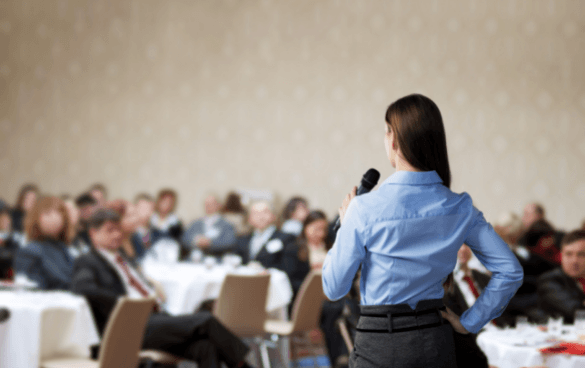 A woman with her back to the camera speaks into a microphone in front of an audience seated at round tables in a conference room.