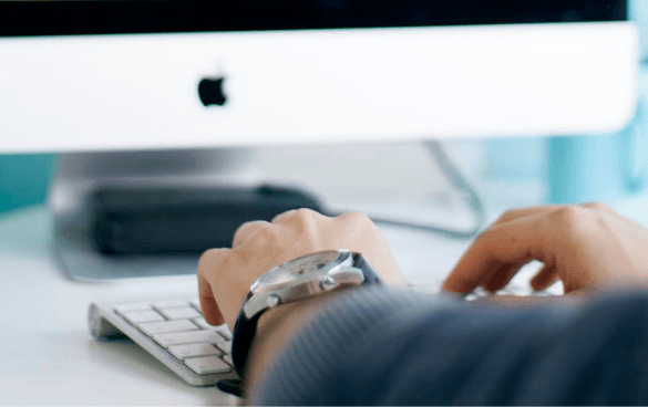 Close-up of a person's hands typing on an Apple keyboard with an iMac and external hard drive in the background.