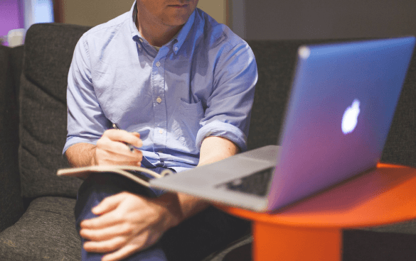 A person in a blue shirt sitting on a couch working on a MacBook and taking notes in a notebook, with a small orange table beside them.