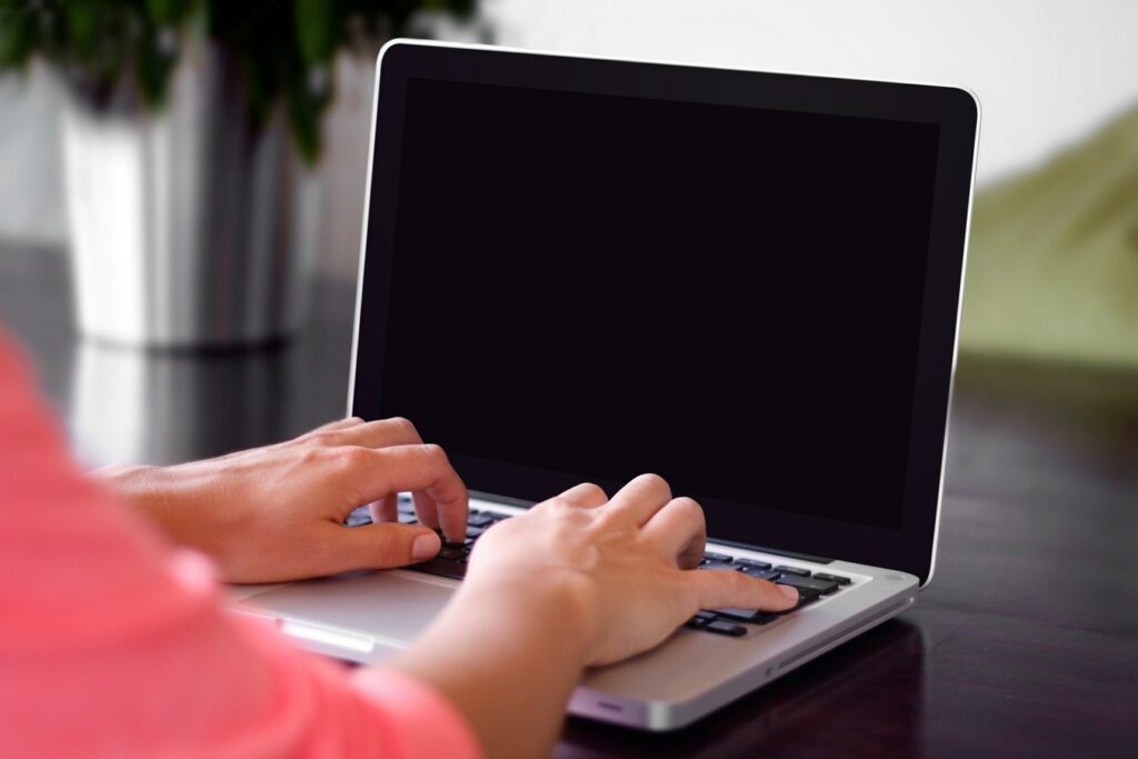 Person in a pink shirt typing on a laptop with a blank screen, set on a dark surface with an indoor blurred background and a hint of greenery.