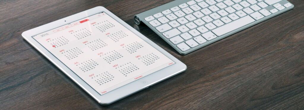 A tablet with a 2015 calendar on the screen next to a white wireless keyboard on a wooden desk.