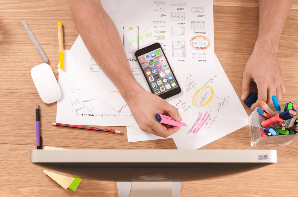 A person's hands are marking a paper with a pink highlighter at a desk cluttered with design sketches, a smartphone, writing utensils, and a computer monitor.