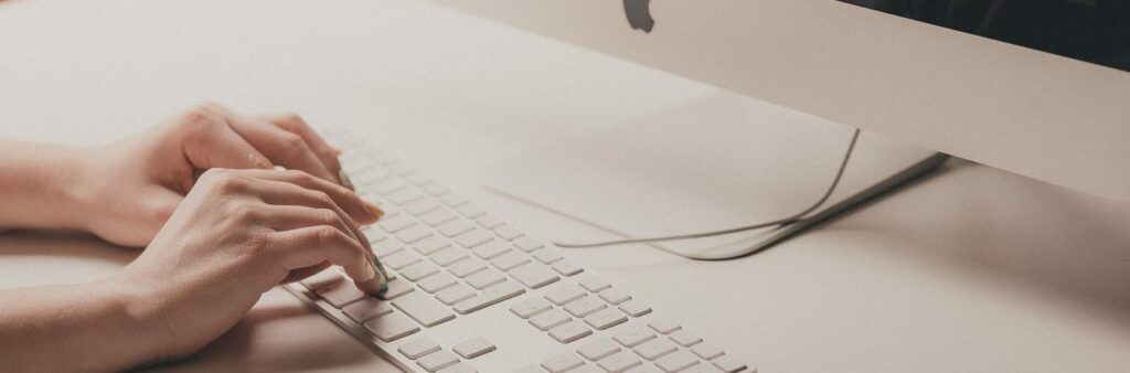 Close-up of hands typing on an Apple keyboard with an iMac in the background.