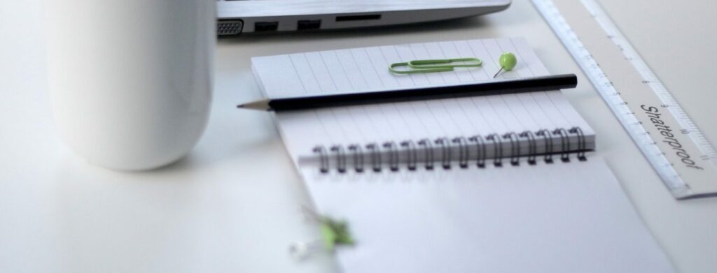 A white desk with a laptop, a white computer mouse, a spiral notebook with a black pen, a green paperclip, a green pushpin, and a clear 'Shatterproof' ruler.