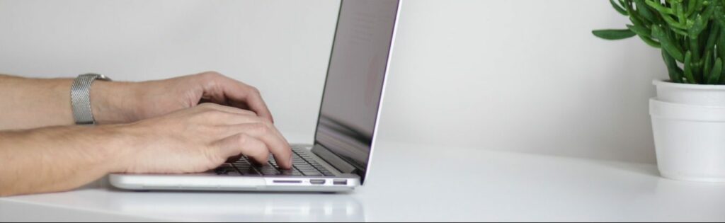 Person's hands typing on a laptop with a silver watch on the left wrist, next to a small potted plant on a white surface with a plain white background.