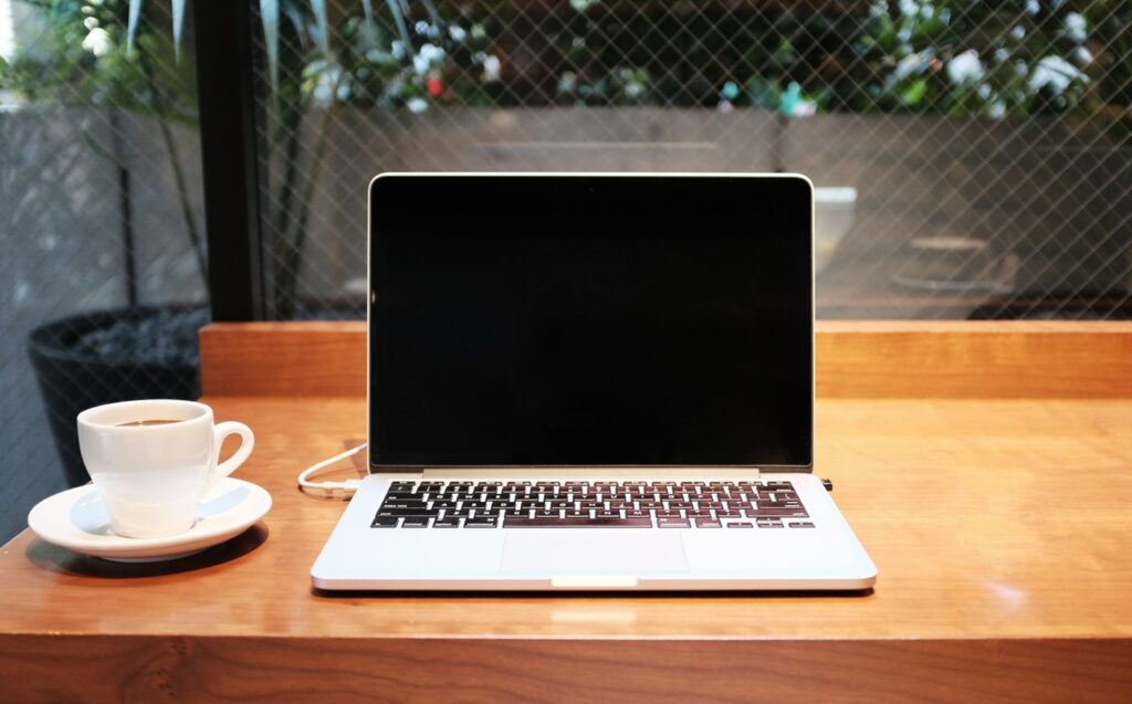 A laptop with a black screen and a cup of coffee on a wooden table, with a window view of greenery in the background.