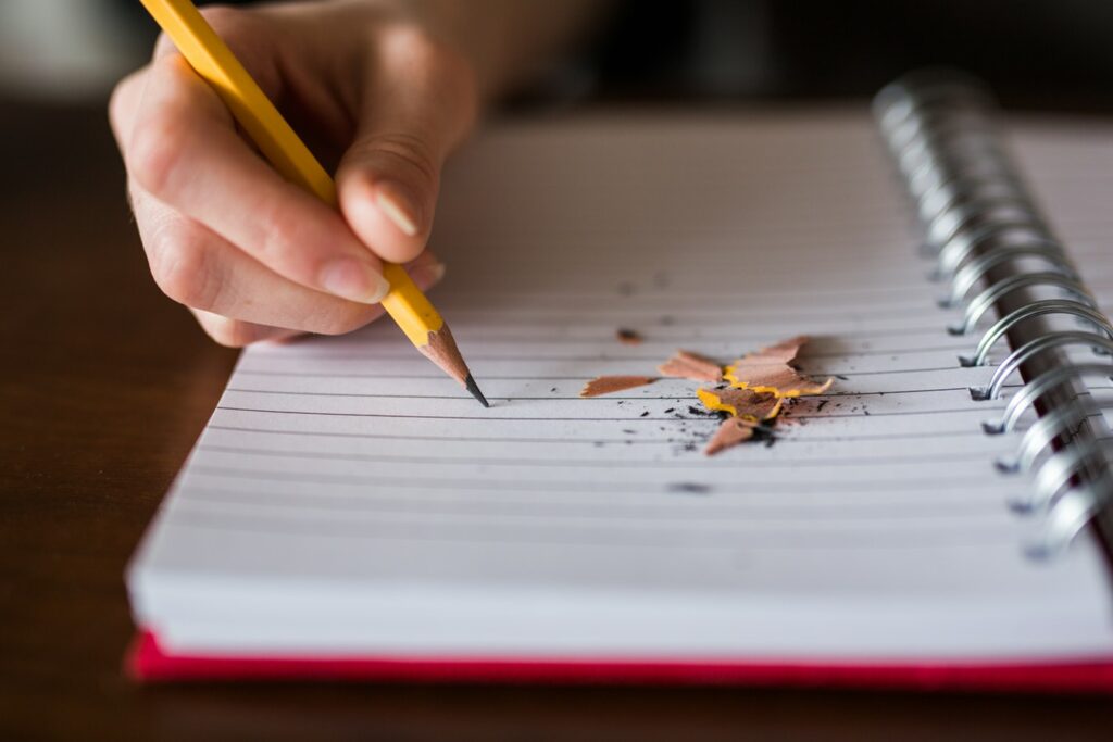 Close-up of a hand holding a sharpened yellow pencil over a lined spiral notebook with pencil shavings on the page.