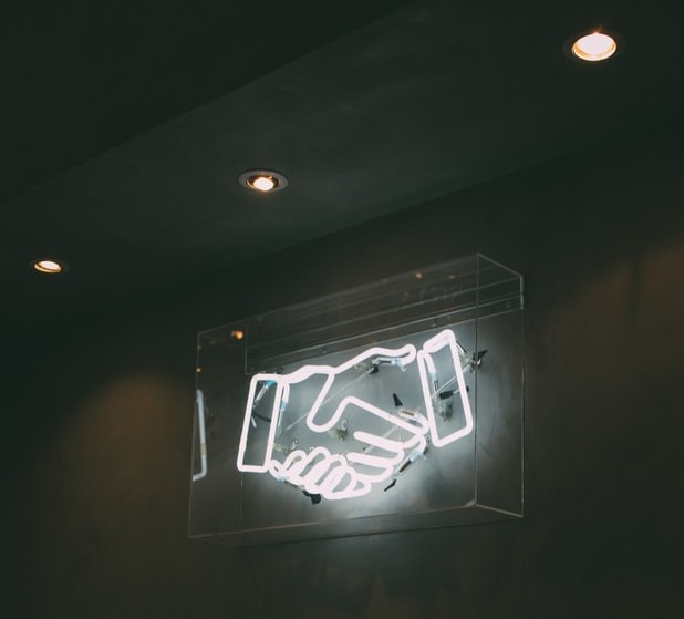 Neon sign of two hands shaking, mounted on a clear backing against a dark wall with warm overhead lighting.