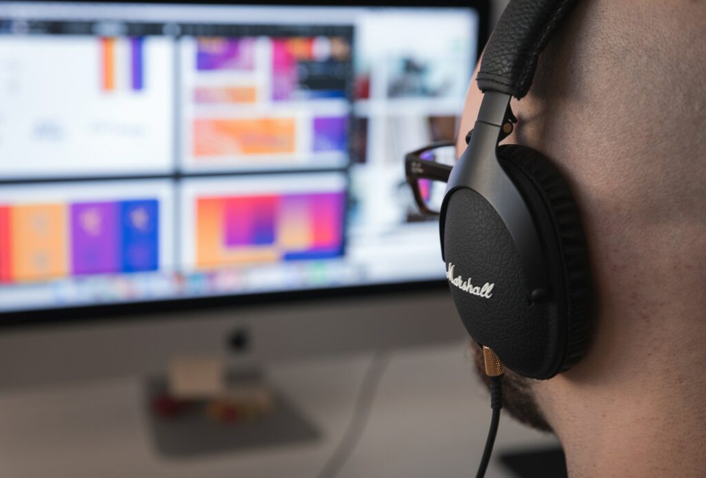 Close-up of a person wearing black Marshall headphones with a blurred computer monitor displaying colorful graphics in the background.