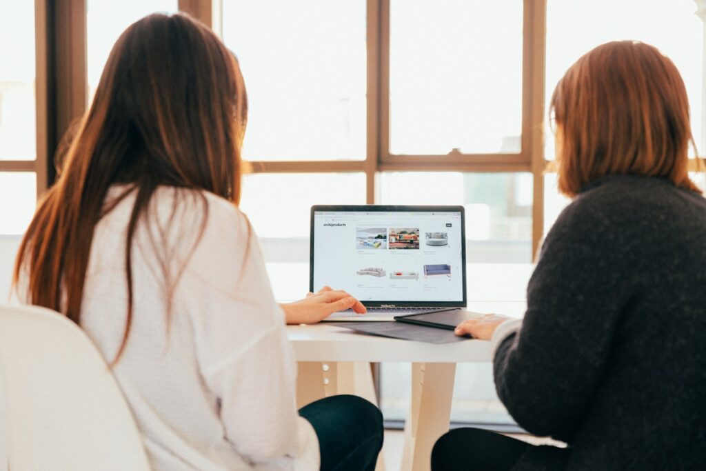 Two women sitting at a table working on a laptop with a website on the screen in a brightly lit room.