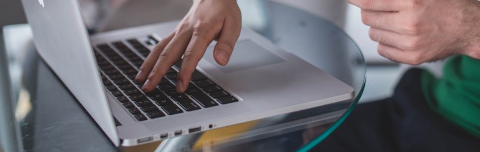 Close-up of a person's hands typing on a laptop keyboard on a glass table, with the lower part of their face visible, wearing a green long-sleeve top.