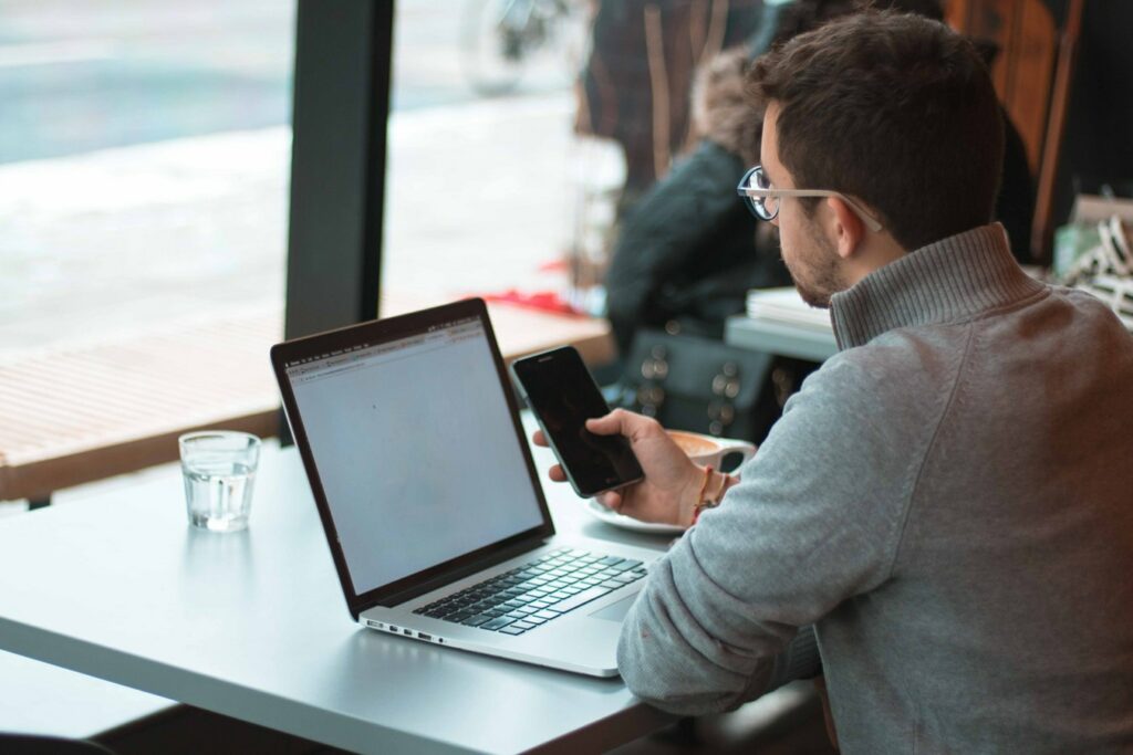 A man in a grey sweater and glasses multitasking with a laptop and smartphone at a cafe table, with a glass of water beside him and a blurred indoor background.