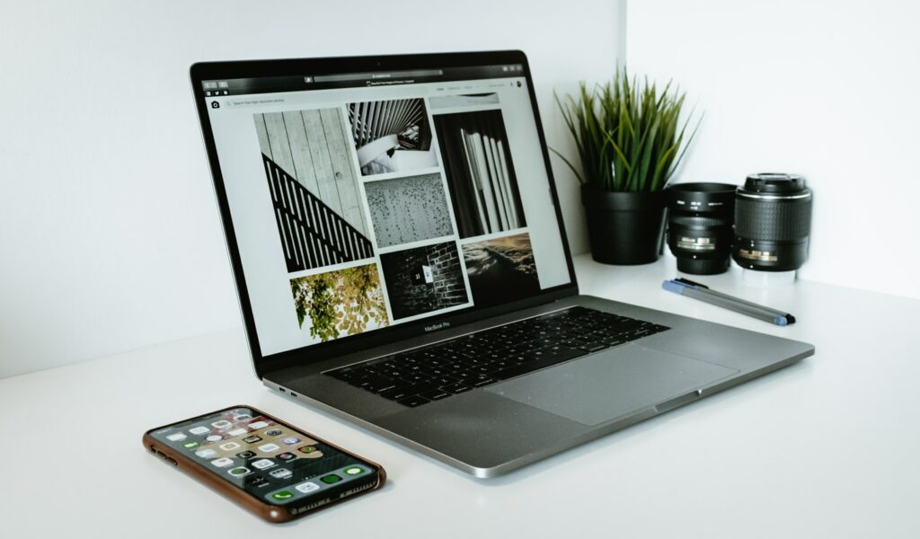 An open laptop displaying black and white photos on a white desk, with a smartphone in front, camera lenses and a pen to the right, and a potted plant behind it.