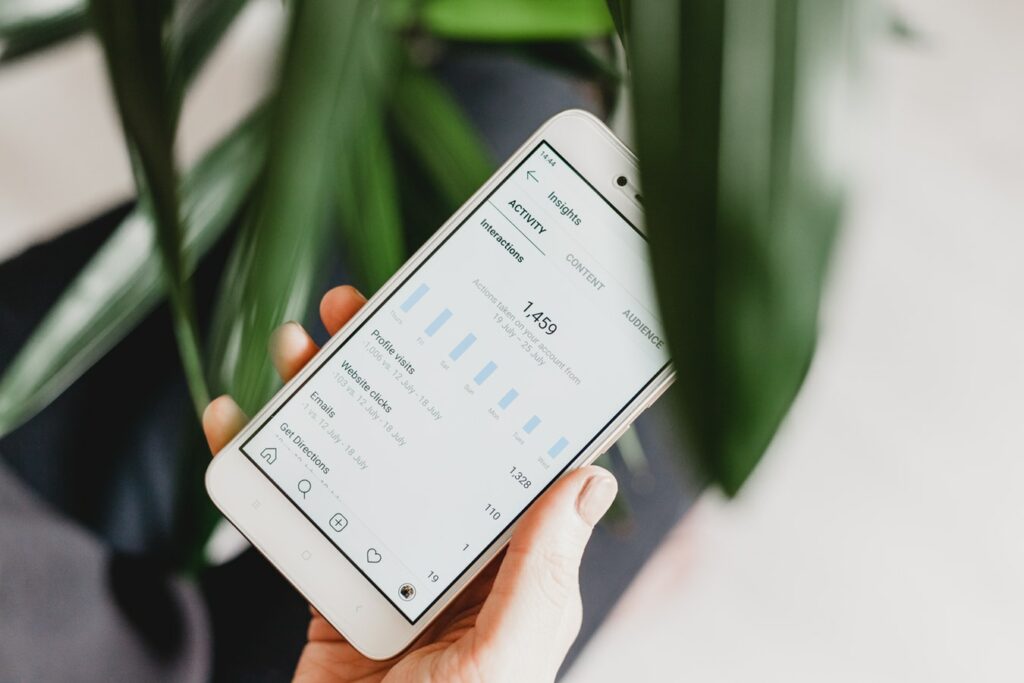 Person holding a smartphone displaying a social media analytics tool with a bar graph showing interactions, phone visits, and website clicks, with engagement icons below, against a backdrop of a green plant.