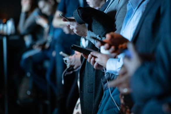 A row of people standing and using their smartphones, with focus on their hands and devices, in an indoor setting.