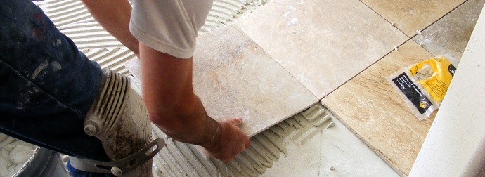 A person installing a ceramic floor tile onto adhesive with knee pads on and a trowel and tile spacers nearby.