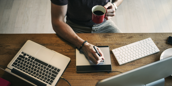 Person at a wooden desk holding a red mug and using a stylus on a digital drawing tablet, with a laptop, wireless keyboard, and part of a desktop monitor visible.