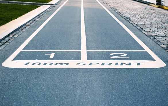 Two lanes of a blue running track marked with '1' and '2' for a 100-meter sprint, bordered by grass and a cobblestone path.