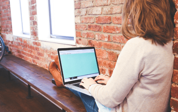 Person sitting on a bench using a laptop with a teal screen in front of a brick wall.