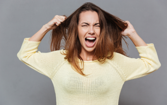 Woman in yellow sweater pulling her hair with a frustrated expression against a grey background.