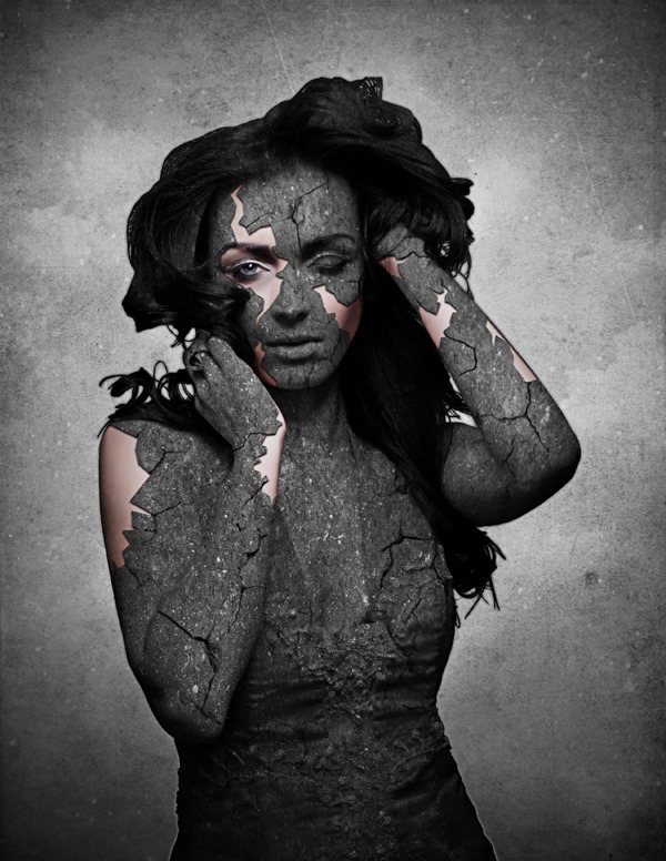 Artistic image of a woman with a cracked stone texture applied to her skin, with reddish layers visible beneath the cracks, hands on her head in a distressed pose against a gray background.