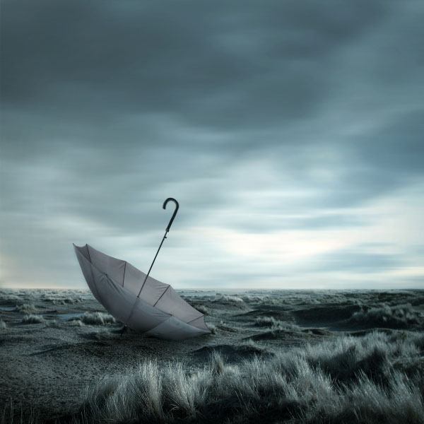 An open umbrella on its side in a desolate, grassy landscape under a cloudy sky with a cool blue-gray color tone.